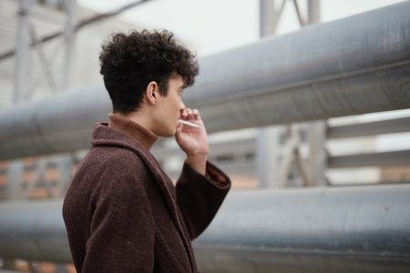 Fashionable young man in a brown coat smoking a cigarette on the side of the street in front of a metal pipe and railingsの写真素材