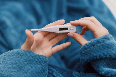 Person holding electronic thermometer in one hand, wrapped in blue blanket, and holding cup of hot tea in other handの写真素材