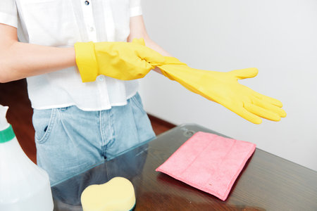 Woman with yellow gloves holding cleaning supplies next to a table with a glass of water and hand sanitizer bottleの写真素材