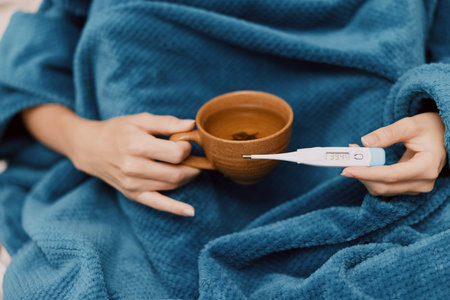 Sick woman wrapped in blue blanket holding thermometer and cup of coffee with thermometer in mouth