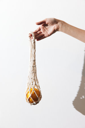 Person holding white macrame bag with orange inside, against white wall, with hand in frontの写真素材
