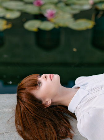 Serene redhead woman laying by pond with water lilies in background capturing a moment of tranquilityの写真素材