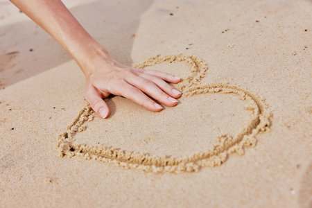 Hand drawing heart in sand on beach with other hand shaping heartの写真素材