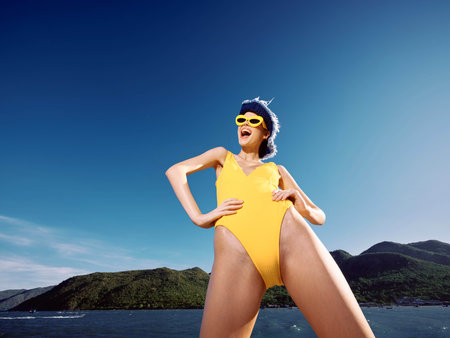 A woman in a yellow swimsuit stands gracefully on the surface of a serene body of water, framed by majestic mountains in the background under a clear, sunny skyの写真素材