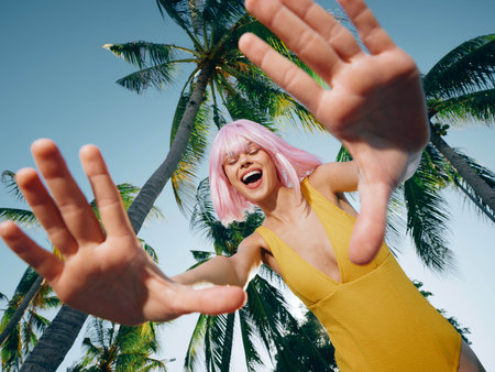 A woman wearing a yellow swimsuit, with her hands raised, stands in front of lush palm trees, capturing a tropical and carefree summer vibeの写真素材