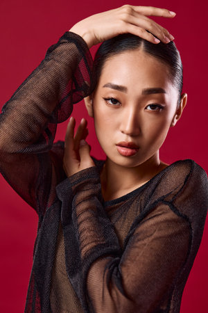 Asian woman in sheer black top posing with hands on head and eyes closed against red background