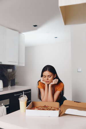Young woman in casual clothing sitting in front of a halfeaten pizza box with drink, contemplating her next sliceの写真素材