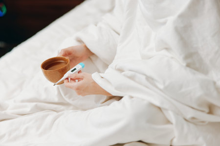 Person sitting in bed with coffee cup and thermometer, wrapped in blanket