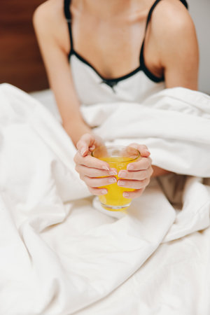 Woman holding a glass of orange juice in bed with white sheet covering her body in the backgroundの写真素材