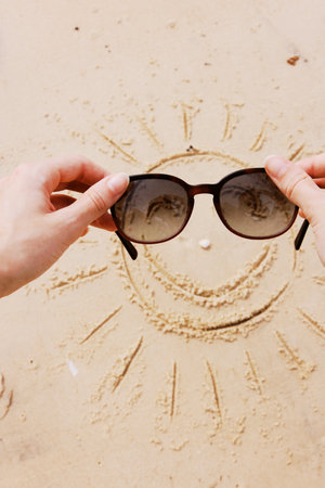 Hands holding sunglasses with smiley face drawn in sand in front of person wearing sunglasses on beachの写真素材