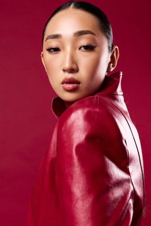 Fashion asian woman in red leather jacket posing against red background with hair in ponytailの写真素材