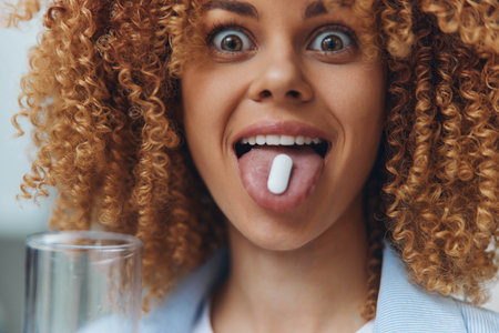 Curlyhaired woman playfully sticking out tongue while holding pill in front of her, concept of healthcare and wellnessの写真素材