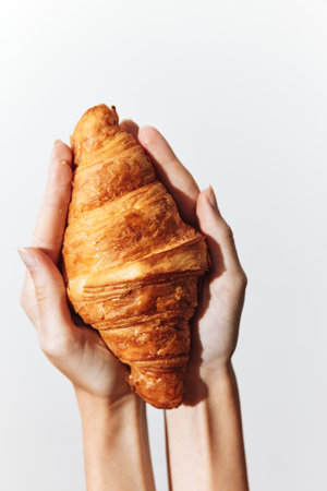 Person holding a croissant in one hand and the other hand holding the croissant over a white backgroundの写真素材