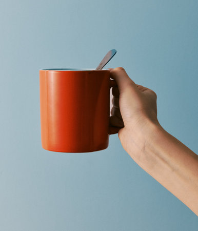 Hand holding an orange coffee mug with a spoon on a blue background, with copy space for text