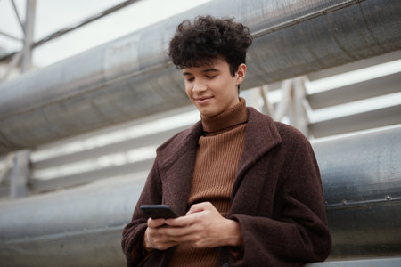 Fashionable young man in brown sweater looking at his cell phone in front of metal pipes