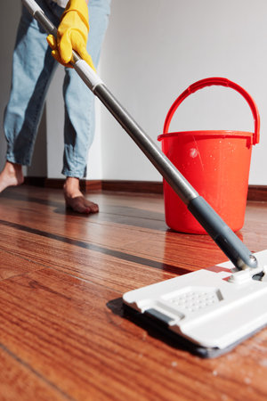 Woman cleaning hardwood floor with mop and bucket of water nearby