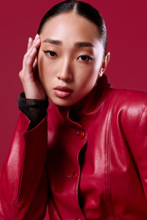 Fashionable asian brunette woman in a red leather jacket posing with her hand on her head and eyes closedの写真素材