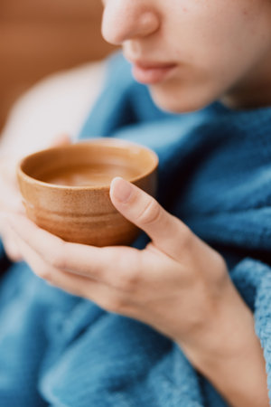 Woman in blue blanket holding bowl of hot water, sitting on bed with cup of teaの写真素材