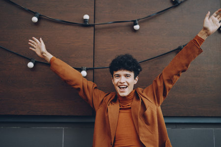 A joyful young man in a brown outfit, raising his arms in excitement against a wooden background with festive lightsの写真素材