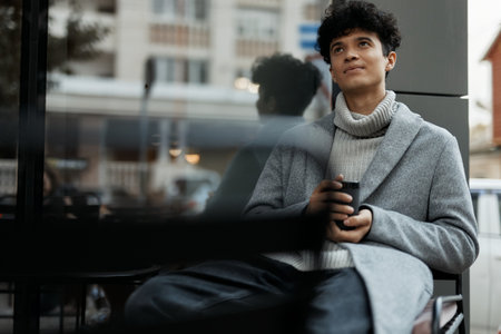 Fashionable young man sitting in front of a building, holding a coffee cup and a phone in his handsの写真素材