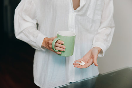 Woman holding a cup of coffee and a pill, standing in front of table with glass of waterの写真素材