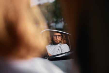 A man with long hair and glasses is seen in a car side mirror, reflecting a thoughtful expression...