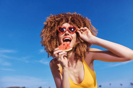Cheerful woman with curly hair wearing heart shaped sunglasses holding a slice of watermelon against a bright blue skyの写真素材