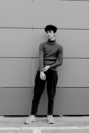 Fashionable young man in black and white attire standing in front of a gray wall