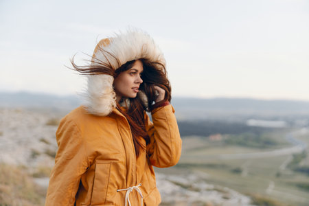 Woman in yellow coat talking on cell phone standing on hilltop with scenic view in travel modeの写真素材