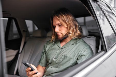 Man sitting in a car, using a smartphone, wearing a green shirt, with long hair, showing a focused...