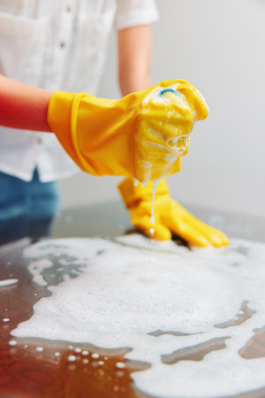 Cleaning woman in yellow rubber gloves wiping table with sponge and soap on white surface