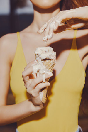 A young woman holding an ice cream cone with melting ice cream on her hands She is wearing a bright yellow tank top The background is softly blurredの写真素材