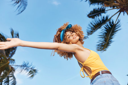 Young woman with curly hair joyfully spreading her arms under a bright blue sky Tropical palm trees frame the scene, creating a lively atmosphereの写真素材