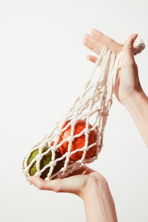 Person holding white net bag with green and red apples, white background, copy space