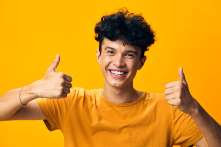 Happy young man showing thumbs up on bright yellow background, smiling joyfully, representing positivity and success with cheerful expression Perfect for uplifting themes and social mediaの写真素材