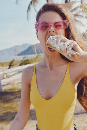 Young woman enjoying ice cream on a tropical beach in a yellow swimsuit Sunlight shines through palm...