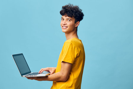 Young man holding a laptop, smiling, wearing a yellow t shirt, against a light blue background, modern and casual concept of technology and youth empowermentの写真素材