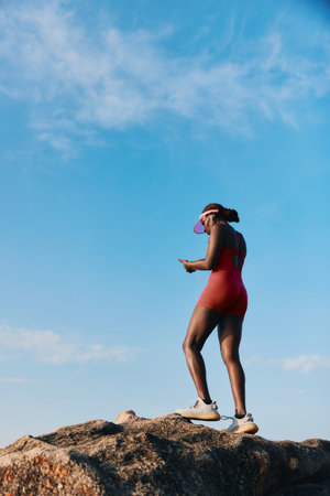Woman in a red swimsuit standing on a rock, looking at her phone against a clear blue sky with wispy cloudsの写真素材