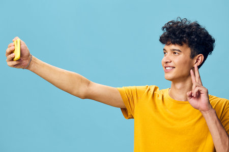 Young man taking a selfie with a smartphone against a bright blue background, wearing a yellow shirt and showing a peace sign with his fingersの写真素材