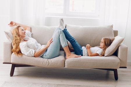 Mother and daughter relaxing on a cozy couch in a bright living room with feet up and smilingの写真素材
