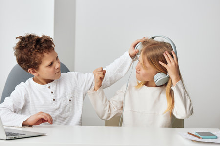 Happy Caucasian children studying at home using laptops and headphones, playing educational games online They are sitting at a table in the cozy living room, focused and engaged in their elearningの写真素材