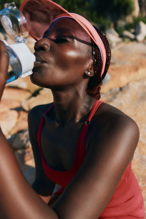 A woman drinking water on a sunny day, wearing a red athletic top and a pink cap Background features rocky terrain, emphasizing an outdoor fitness themeの写真素材