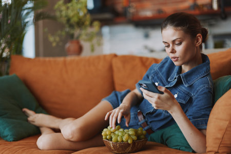 Young woman relaxing on couch with bowl of grapes and cell phone, enjoying a quiet moment at homeの写真素材