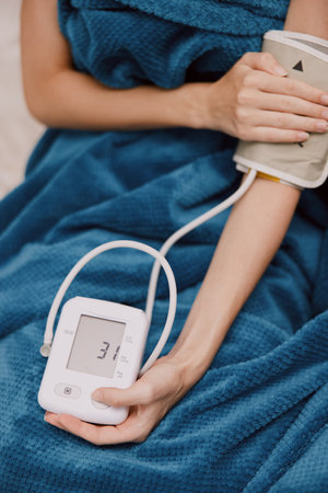 Woman in blue blanket sits on bed holding blood pressure monitor and wearing cuff on armの写真素材