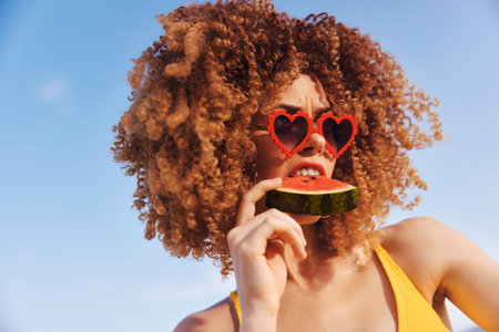 Woman with curly hair wearing heart shaped sunglasses, holding a piece of watermelon against a bright blue skyの写真素材
