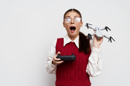 Excited girl holding a drone and remote controller in front of a light background Her glasses reflect amazement as she engages with technology enthusiasticallyの写真素材