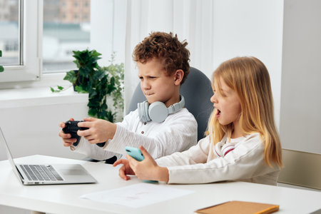 Happy Caucasian school children studying and playing at home using laptops and tablets for online elearning With concentration and joy, the boy and girl sit at a table in their living room, wearingの写真素材