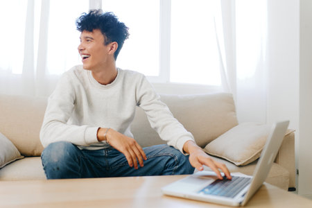 Smiling young man working on laptop at home, modern and cozy interior with natural light and soft colors, relaxed atmosphere, studying or remote work conceptの写真素材