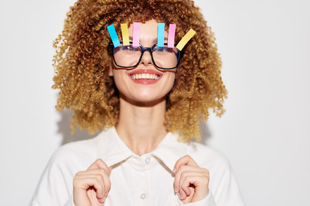 Creative woman with curly hair and colorful clips in her glasses, smiling happily against a bright white background showcasing playful personality and humorの写真素材