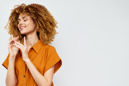cheerful young woman with curly hair in an orange shirt, joyfully expressing happiness against a...